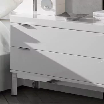 The Modern Miami Style Large White Bedside Table with two drawers, adorned with sleek metal handles and shiny chrome legs, stands adjacent to a white bed dressed in white linens. A circular object and a decorative dish sit atop the well-lit and minimalistic bedside table.