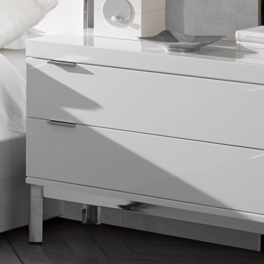 Modern Miami Style Large White Bedside Table The Modern Miami Style Large White Bedside Table with two drawers, adorned with sleek metal handles and shiny chrome legs, stands adjacent to a white bed dressed in white linens. A circular object and a decorative dish sit atop the well-lit and minimalistic bedside table.