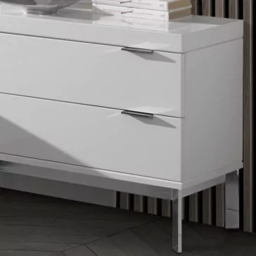 The **Modern Miami Style Large White Bedside Table** displays a contemporary aesthetic with its two sleek drawers, each adorned with minimalist chrome handles. It stands on slim, polished metal legs and is positioned against a textured wall. The surface of the dresser holds a few books and a decorative item.