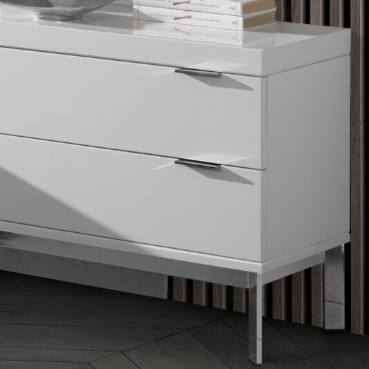 Modern Miami Style Large White Bedside Table The **Modern Miami Style Large White Bedside Table** displays a contemporary aesthetic with its two sleek drawers, each adorned with minimalist chrome handles. It stands on slim, polished metal legs and is positioned against a textured wall. The surface of the dresser holds a few books and a decorative item.