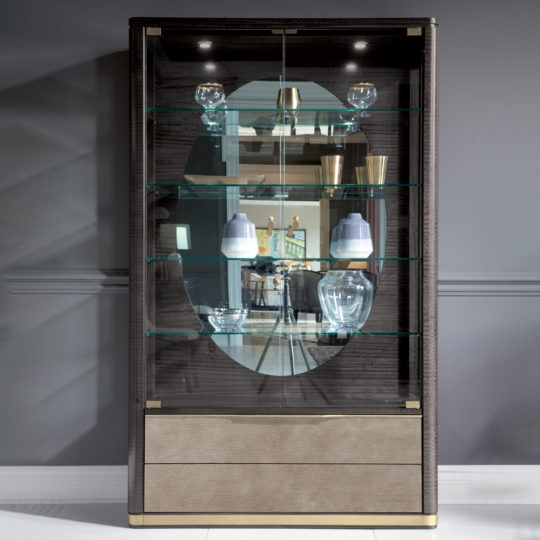 The Modern Leather Front Display Cabinet showcases a sleek design with curved edges, featuring shelves that elegantly display glassware such as a decanter and bowls. It includes backlighting to enhance the presentation and offers additional closed storage at the bottom. The mirrored back panel reflects the surrounding room, adding depth and sophistication to the piece.