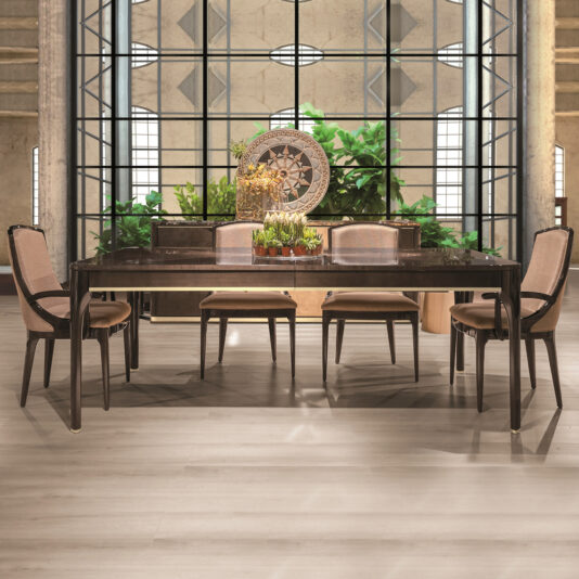 The Modern Maple Dining Table Set showcases a sleek dark wooden table accompanied by six upholstered chairs. Positioned behind the table, a large industrial-style window provides views of lush green plants. The background is enhanced with a decorative centerpiece and a large circular wall ornament.