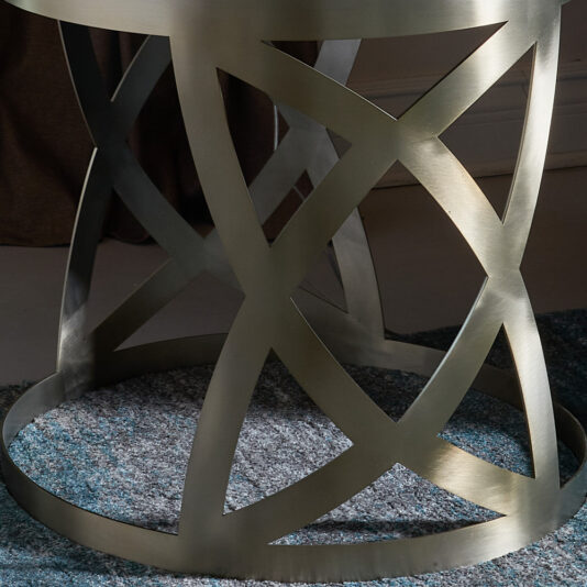 A close-up image of the Modern Dark Walnut Occasional Table base, crafted from smooth and reflective metal with an intricate crisscross design, resting on a blue and gray textured rug. The base features intersecting, curved lines.