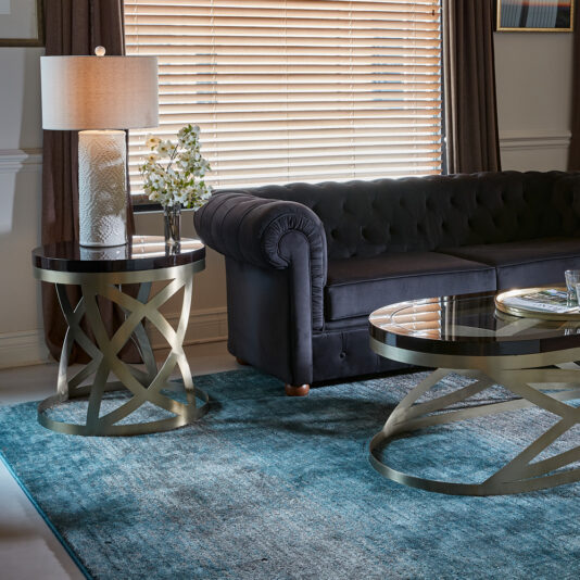 A modern living room showcases a plush, dark-colored sofa with a button-tufted back. The room includes both a glass-top coffee table and side table featuring gold crisscross metal bases, which pair perfectly with the Modern Dark Walnut Occasional Table. A beige lamp and a vase filled with white flowers adorn the side table, while a teal-blue rug and window blinds pull the whole space together.