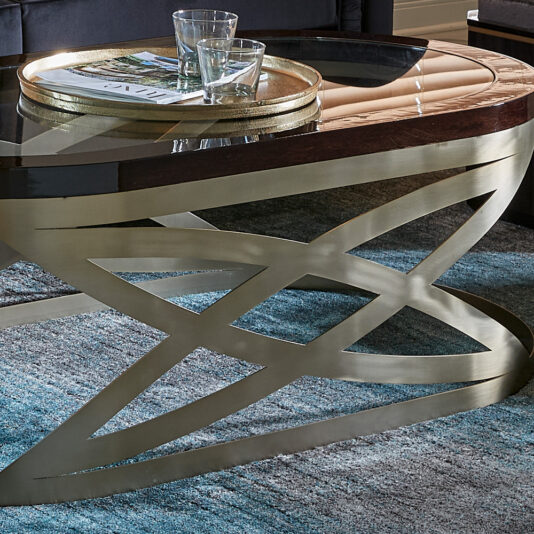 Modern Oval Coffee Table A sleek Modern Oval Coffee Table with a refined glass top and an ornate gold metal base rests on a blue patterned rug. The stylish coffee table holds two glass tumblers and a tray carrying a magazine. In the background, part of a dark sofa is visible.