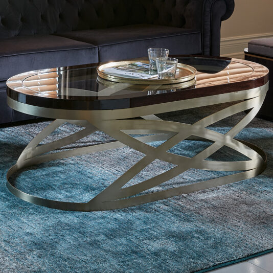 The Modern Oval Coffee Table, featuring an intricate metallic base and a gleaming glass top, is placed on a teal area rug. The table artfully holds a round tray with a stack of magazines and two glasses, set against the backdrop of a dark, tufted sofa.