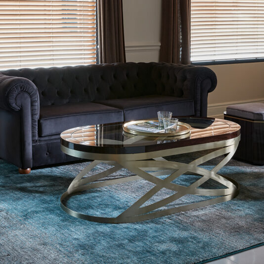 Modern Oval Coffee Table A modern living room featuring a dark, tufted couch, the Modern Oval Coffee Table with a glass top and intricate gold base, and a teal area rug. Nearby is a small ottoman, two glasses, and a magazine on the table. Blinds cover the windows in the background.