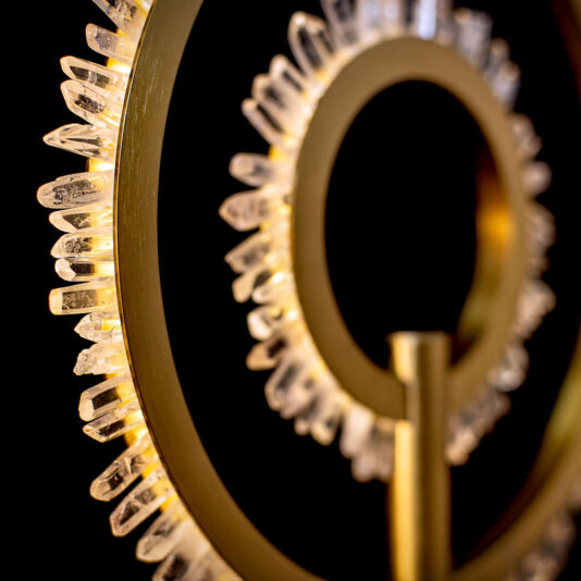 Close-up of the Modern Quartz Crystal Hoop Table Lamp, featuring a circular, gold-colored light fixture adorned with clear, crystal-like accents on its outer edge. Set against a dark background, the intricate details of the crystals and the metallic finish are beautifully highlighted.