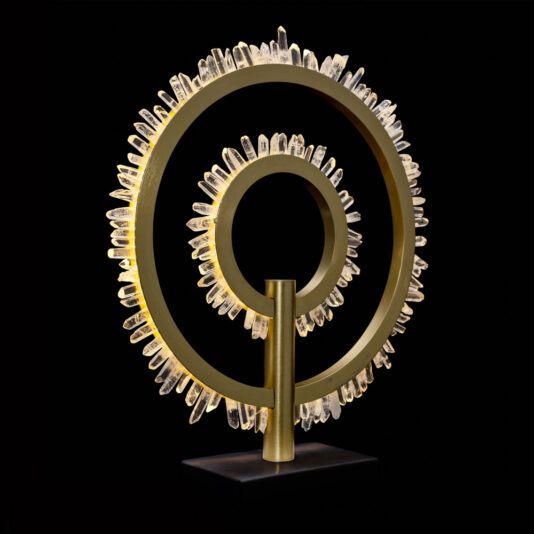 The Modern Quartz Crystal Hoop Table Lamp consists of two intertwined golden rings adorned with cascading clear crystals against a black backdrop, creating a visually striking and elegant appearance.
