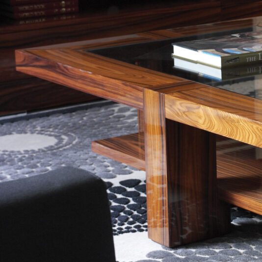 Modern Rectangular Coffee Table With Glass Top The Modern Rectangular Coffee Table with Glass Top is positioned on a dark, textured rug featuring circular patterns. Several books are neatly stacked on the table. The corner of a black sofa is partially visible in the foreground, and a wooden bookshelf can be seen in the background.