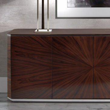 The Modern Rosewood Sideboard, boasting a rich, dark wood finish and adorned with a striking sunburst pattern on its cabinet doors, is set against a light gray textured wall. Above it rests a sleek geometric lamp and a framed abstract painting, while the floor beneath is polished and light-colored.