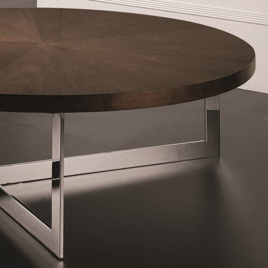 The Modern Round Coffee Table, featuring a dark wooden top and a polished chrome geometric base, sits elegantly against a plain white wall, perfectly complementing the dark-colored floor.