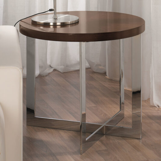 The Contemporary Round Side Table, featuring a brown wooden top and shiny silver metal legs in a geometric design, stands proudly on a wooden floor beside a white couch. Adorned with a lamp, it effortlessly complements the white curtains hanging in the background.