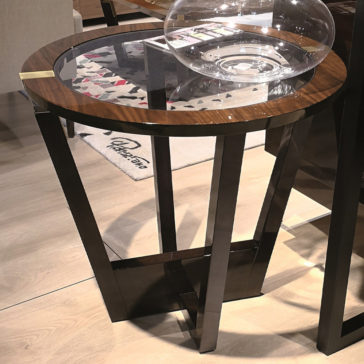 Modern Round Glass Top Occasional Table The Modern Round Glass Top Occasional Table, which boasts a wooden top and metal legs with a round glass insert in the center, is adorned with a clear, empty glass bowl. This contemporary piece is placed on a light wood floor adjacent to a patterned rug.