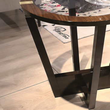 Modern Round Glass Top Occasional Table The Modern Round Glass Top Occasional Table, featuring wood and metal legs, stands on a hardwood floor. A part of a patterned rug is visible underneath it, while its glass top reflects a section of the nearby sofa.