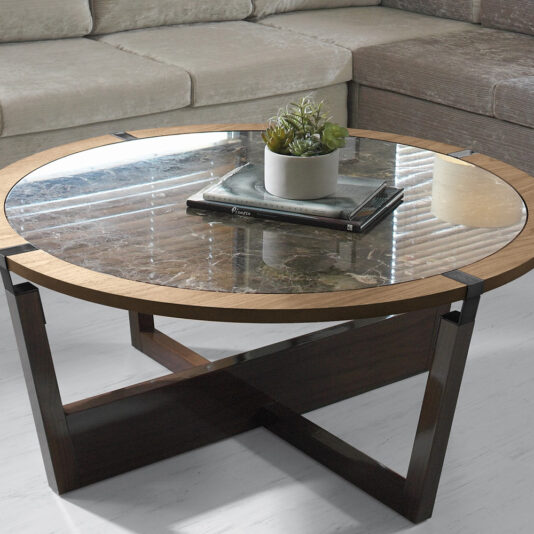 In the center of a living room sits the Modern Round Marble Coffee Table, featuring a glass top and dark wooden base and supports. A potted plant and a book rest on the table, which stands on a light-colored floor with a beige sectional sofa in the background.