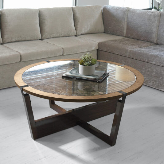 A modern living room showcasing the Modern Round Marble Coffee Table, which features a sleek marble top with a wooden rim and dark geometric legs. The table displays a small potted succulent arrangement and two closed books. In the background, there is a comfortable light grey sectional sofa.