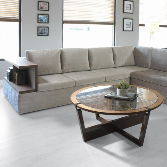 A contemporary living room showcasing a beige sectional sofa paired with a dark wood side table. The space boasts a Modern Round Marble Coffee Table edged in light wood, complemented by a small potted plant and books. Large windows with blinds and framed pictures adorn the walls, completing the stylish look.