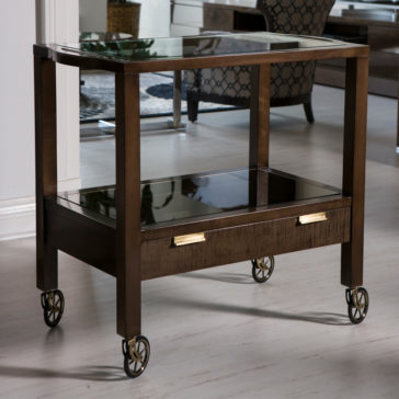 Modern Trolley With Glass Top The Modern Trolley With Glass Top boasts a stylish rectangular wooden bar cart design with a sleek glass middle shelf and a convenient lower drawer adorned with two gold handles. Supported by four sturdy legs equipped with elegant gold caster wheels, this trolley perfectly complements the wooden flooring and contemporary furniture in the background.