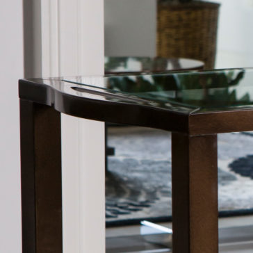 Modern Trolley With Glass Top A close-up view of the Modern Trolley With Glass Top, featuring a sleek dark wooden frame, situated in an indoor space. The background reveals a softly blurred room with a patterned rug and a wicker basket.