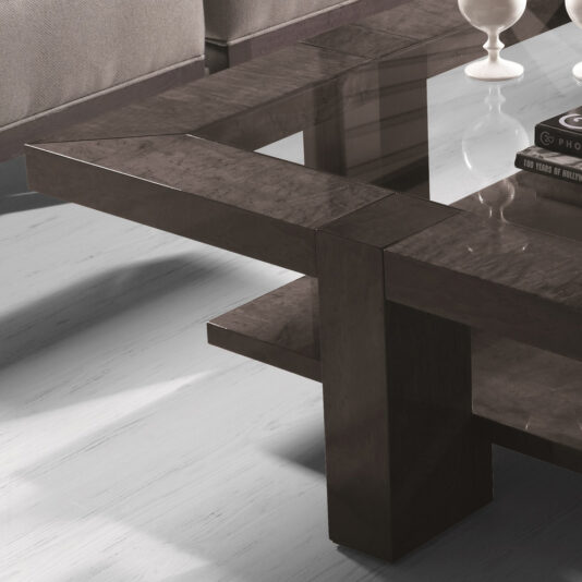 The Modern Square Coffee Table With Glass Top boasts a sleek, geometric design with a glass top and dark wood accents, creating a perfect contrast against the light floor. In the background, part of a gray sofa and two white decorative objects further enhance the contemporary decor.