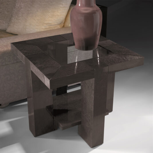The Modern Square Side Table With Glass Top, characterized by its dark brown wood and geometric design, is placed next to a cream-colored sofa, combining style and functionality with its convenient lower shelf. In the dimly lit background creating a cozy atmosphere, the table is adorned with a decorative reddish-brown vase.