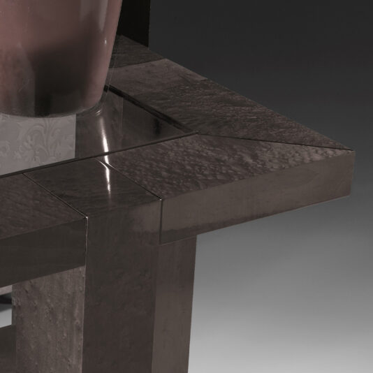 Close-up of the Modern Square Side Table With Glass Top. The table boasts a sleek, glossy surface with a dark finish and features a geometric design characterized by sharp edges and clean lines. Part of a reflective vase is visible on the glass top. The background transitions from dark to light gray in a gradient pattern.