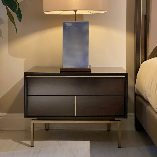 The Modern Stylish Bedside Table with two drawers stands on a beige-colored floor. On top, there is a large rectangular blue lamp with a beige lampshade, casting a warm light. Part of a bed frame and a green plant are partially visible in the background.