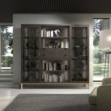 Modern Stylish Wooden Bookcase A contemporary living room showcases the Modern Stylish Wooden Bookcase with glass doors, elegantly filled with books and decorative items. A beige sofa and a large floor lamp are also visible. Natural light pours in through the windows, highlighting the lush green foliage outside.