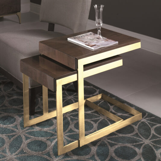 Modern Occasional Table Nest The Modern Occasional Table Nest features two rectangular side tables with elegant gold metal frames and dark wooden tops. The larger table is utilized to hold a magazine and a partially filled glass of liquid. Both tables are situated on a patterned rug, complementing the room’s decor with a gray armchair in the background.