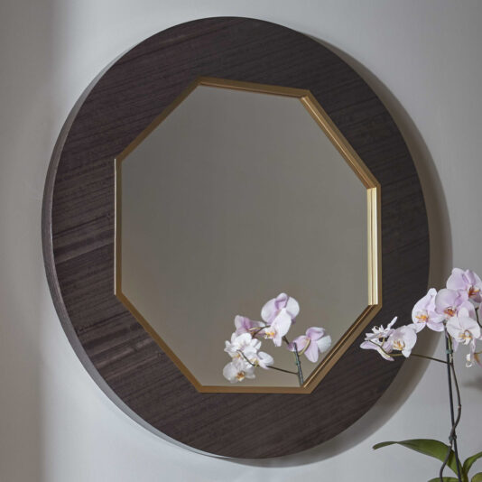 The Modern Wooden Octagonal Mirror showcases a gold-colored inner frame. Reflected in the mirror is a part of the wall along with some light-colored orchids in bloom, artfully placed on the lower right side.