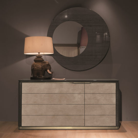 A dimly lit room features the Modern Wooden Dresser And Mirror Set, showcasing a sleek dark surface complemented by light-colored drawers. Atop the dresser is an ornate lamp with an elephant base, casting a warm glow through its beige lampshade. Above the dresser hangs a large round mirror with a dark frame, adding to the contemporary aesthetic.