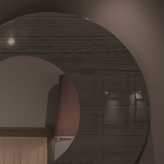 A close-up of a wall-mounted circular mirror from the Modern Wooden Dresser And Mirror Set slightly reflecting a dimly lit room. The mirror features a dark, textured frame. A portion of a wooden headboard and burgundy lampshade are visible in the reflection against the grey background wall.