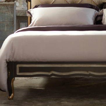 The Ornate Button Upholstered Italian Bed features a meticulously made bed with crisp white linens and rich dark purple accents, complemented by a beige, ornately designed headboard. The dark wood frame is adorned with decorative carvings that enhance the Italian aesthetic. Adding to the room's rustic charm, a vintage dresser can be seen in the background.