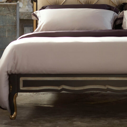 Ornate Button Upholstered Italian Bed The Ornate Button Upholstered Italian Bed features a meticulously made bed with crisp white linens and rich dark purple accents, complemented by a beige, ornately designed headboard. The dark wood frame is adorned with decorative carvings that enhance the Italian aesthetic. Adding to the room's rustic charm, a vintage dresser can be seen in the background.