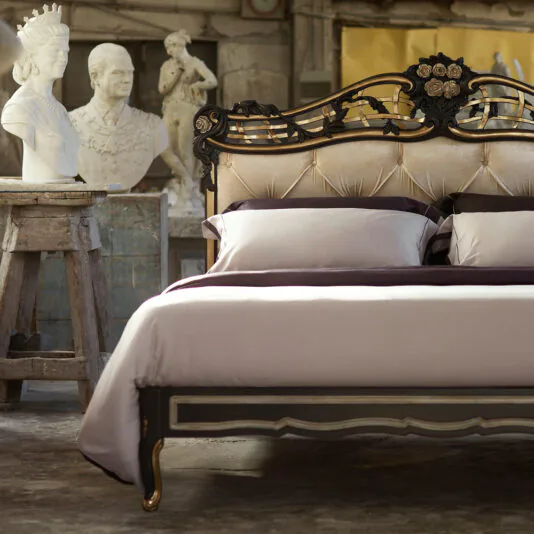 Ornate Button Upholstered Italian Bed A luxurious bedroom features an Ornate Button Upholstered Italian Bed adorned with plush bedding. In the foreground, a small workbench holds two classical busts, one of a man and one of a woman. The background showcases a rustic wall adorned with artistic decorations.