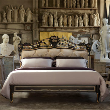 In an art studio filled with classical sculptures and busts, an Ornate Button Upholstered Italian Bed boasting a tufted headboard and intricate design is exquisitely decorated. The bed is adorned with light-colored bedding and situated in a rustic, dimly lit room.