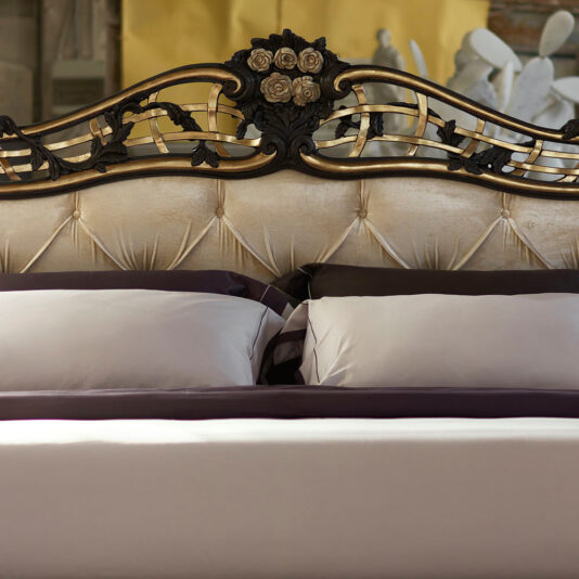 Ornate Button Upholstered Italian Bed The Ornate Button Upholstered Italian Bed is crowned with a luxurious headboard featuring intricate floral designs and gold accents. This elegant bed showcases beige tufted upholstery and is adorned with neatly arranged white and dark-colored pillows and bedding, creating a sophisticated and inviting atmosphere.