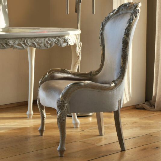 The Ornate Carved Occasional Chair, elegantly adorned with intricate floral designs on its backrest and legs, is upholstered in white fabric and placed beside a round table that exhibits matching decorative carvings. The setting is completed by the polished wooden floor in a warmly lit room.