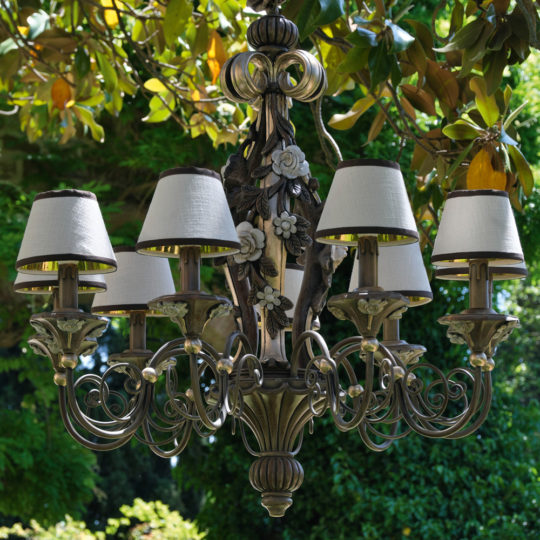 An Ornate Classic Rose Chandelier With Shades, featuring intricate floral and scrollwork designs, hangs outdoors against a background of lush green foliage and trees. The chandelier has a vintage, elegant look with its eight lampshades adding to its grandeur.