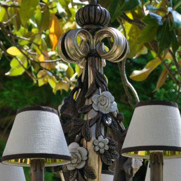 Ornate Classic Rose Chandelier With Shades The Ornate Classic Rose Chandelier With Shades, featuring intricate floral carvings and adorned with three white lampshades, is positioned outdoors against a backdrop of lush green foliage and sunlit leaves.