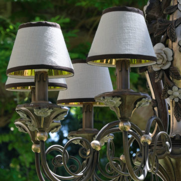 Ornate Classic Rose Chandelier With Shades A close-up of the Ornate Classic Rose Chandelier With Shades reveals its intricately designed metal arms and floral accents. The chandelier features three fabric lampshades with a white exterior and gold interior, contrasting beautifully against a background of lush green foliage.