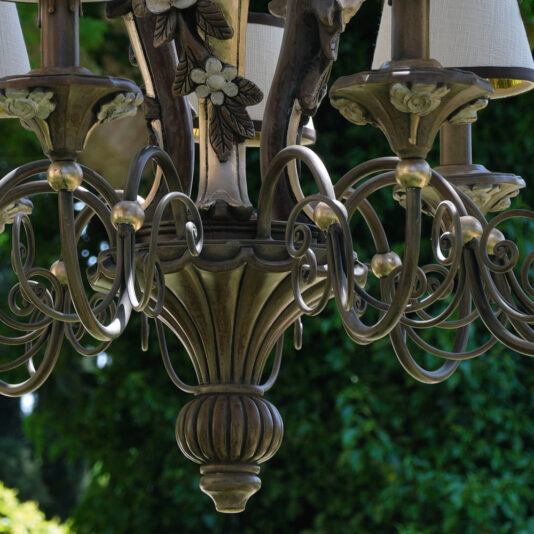 Ornate Classic Rose Chandelier With Shades A close-up of the Ornate Classic Rose Chandelier With Shades showcases its intricate metalwork and floral designs. Several candle-shaped lights, each adorned with delicate shades, are mounted on the chandelier's elaborate arms. In the background, a blurred outdoor scene with lush greenery creates a picturesque setting.