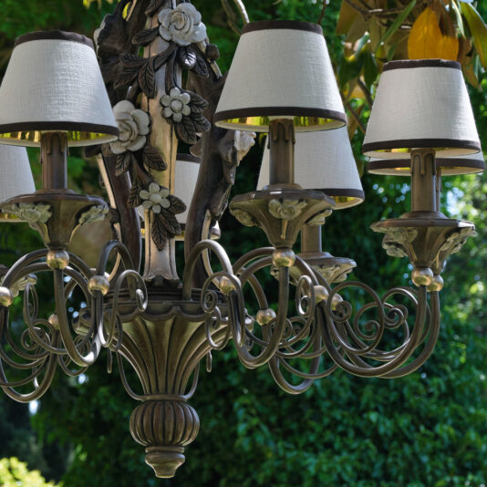 Ornate Classic Rose Chandelier With Shades The Ornate Classic Rose Chandelier With Shades, an elegant vintage-style light fixture adorned with intricate scrollwork and floral details, featuring six candle-shaped fixtures topped with white lampshades, is hanging outdoors against a backdrop of lush, green foliage.