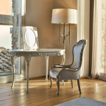 Ornate Dressing Table With Hidden Mirror An Ornate Dressing Table With Hidden Mirror in vintage style, paired with a matching cushioned chair, sits beside a tall floor lamp. The setting is illuminated by natural light streaming through large windows, providing a view of an old building with a tiled roof.