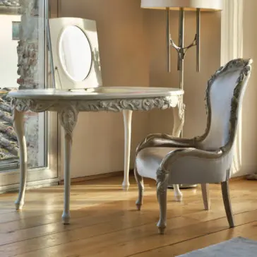 Ornate Dressing Table With Hidden Mirror A stylish vanity setup features the Ornate Dressing Table With Hidden Mirror, paired with a matching chair, both adorned with intricate carvings. The table stands gracefully beneath an oval mirror while a floor lamp with a cylindrical lampshade is placed beside it. Sunlight streams through a nearby window, highlighting the wooden floor.