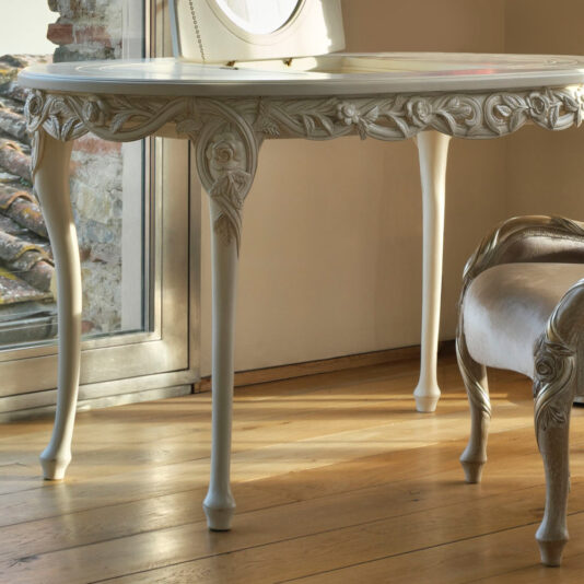 Ornate Dressing Table With Hidden Mirror An Ornate Dressing Table With Hidden Mirror, featuring intricate floral carvings on its sides and legs, stands next to a window. This vintage-style white wooden table is adorned with slender, elegantly curved legs. The sunlight illuminates the polished wooden floor, creating a warm ambiance.