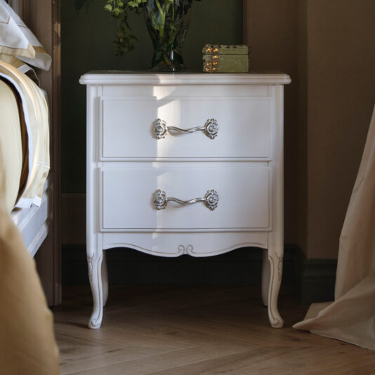 An Ornate Italian Bedside Cabinet in white wood, featuring two drawers with silver handles, stands against a green wall. A small green decorative box and a vase of flowers are placed on top of the cabinet. Nearby is the corner of a bed adorned with white and yellow bedding.