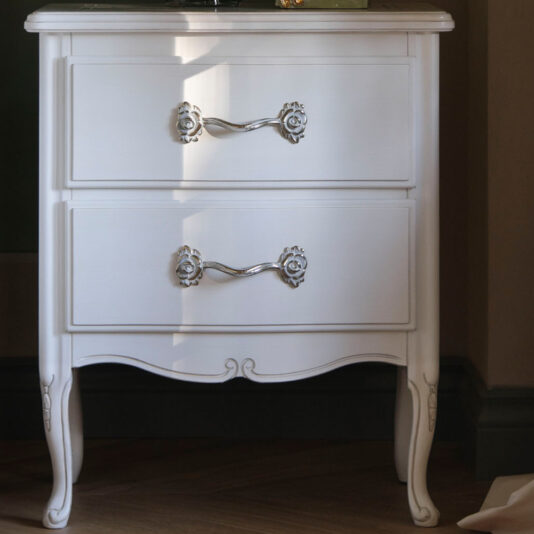 Ornate Italian Bedside Cabinet The Ornate Italian Bedside Cabinet boasts a charming vintage style, finished in white and featuring two drawers adorned with intricate silver handles. It stands on elegant, curved legs and is accented by a decorative trim at the bottom. The top surface of the cabinet is partially visible with a decorative item placed on it, set within an indoor environment.