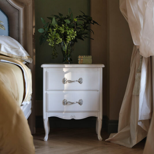 Ornate Italian Bedside Cabinet An Ornate Italian Bedside Cabinet, featuring a white finish with two drawers and intricate handles, is positioned next to a bed adorned with beige and white bedding. On the cabinet sits a green leafy plant in a clear vase and a small decorative item. Curtains frame the right side of the image.